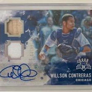 May include: A baseball card featuring Willson Contreras of the Chicago Cubs. The card has a blue and white watercolor background, with a patch of wood and fabric. It includes an autograph and is encased in a protective holder.
