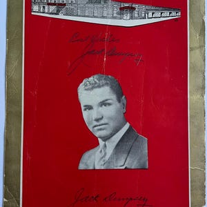 May include: Vintage red and gold-edged promotional item featuring a black and white image of a man in a suit. The image includes a drawing of a building and handwritten text, including the name "Jack Dempsey".