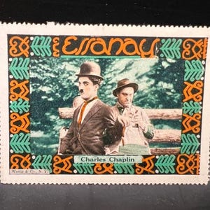 May include: A vintage postage stamp featuring a black and white image of Charlie Chaplin and another man. The stamp has a decorative border with orange, green, and black patterns. The text "Essanay" and "Charles Chaplin" are visible.
