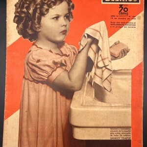 May include: Vintage magazine cover featuring a young girl with curly hair, holding a towel and soap near a sink. The cover has text in Spanish, including "FILMS SELECTOS" and "SHIRLEY TEMPLE". The color palette is primarily peach, red, and cream.