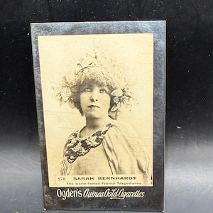 May include: A rectangular vintage cigarette card featuring a sepia-toned portrait of Sarah Bernhardt wearing an ornate headpiece and decorative collar. The text reads 178 SARAH BERNHARDT The world-famed French Tragedienne and Ogden's Guinea Gold Cigarettes.
