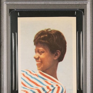 May include: A 1960 Hemmets Journal trading card featuring Wilma Rudolph. The card shows a smiling portrait of Rudolph wearing a red, white, and blue striped top. The card is graded and encased in a protective holder.