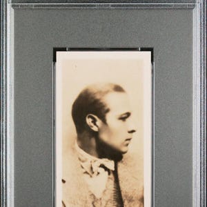 May include: A 1926 Lambert & Butler trading card featuring a sepia-toned profile portrait of Rudolph Valentino. The card is encapsulated in a clear PSA protective slab with a label reading 1926 LAMBERT & BUTLER RUDOLPH VALENTINO #5 EX-MT 6.