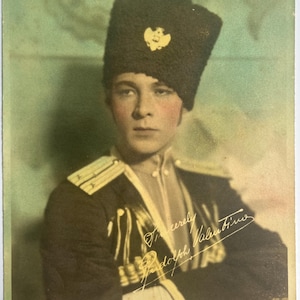 May include: A vintage color photograph of a person wearing a dark military uniform and a tall, dark hat with a gold emblem. The uniform has gold trim and epaulets. The photograph is signed "Sincerely Rudolph Valentino".