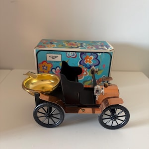 Puede incluir: Un cenicero con forma de coche antiguo, con carrocería color cobre, ruedas negras y un cenicero dorado. El coche tiene un asiento negro y está colocado frente a una caja con estampado floral. Objeto decorativo.