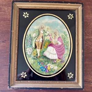 May include: An antique framed embroidery featuring a couple in period clothing in a garden setting. The oval embroidery is set against a black background with a gold frame. The frame has gold accents in the corners.