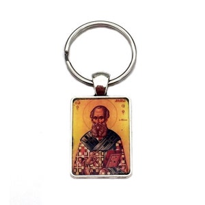 May include: A rectangular keychain featuring a religious icon of a bearded man with a halo, holding a book. The image has a gold background and is framed in silver, attached to a silver key ring.