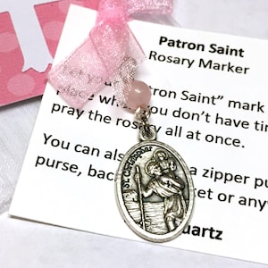 May include: A silver-toned St. Christopher rosary marker with a pink quartz bead and ribbon. The marker is attached to a card with the text "Patron Saint Rosary Marker". A pink polka dot card with a white cross is in the background.