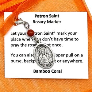 Puede incluir: Un amuleto de plata con una imagen de Santa Filomena. El amuleto está unido a una cuerda de cuentas rojas y blancas. El amuleto está etiquetado como "Patron Saint Rosary Marker" y "Bamboo Coral".