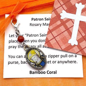 May include: A small silver charm with a colorful image of a saint, a red bead, and a silver clasp. The charm is attached to a white card with the text "Patron Saint Rosary Charm" and instructions on how to use it. The card is on a bright orange background.