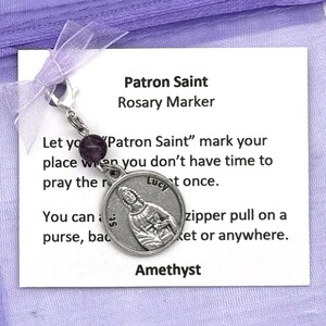 May include: A purple amethyst rosary marker with a silver charm featuring a saint. The charm has the words "St. Lucy" on it. The card reads "Patron Saint Rosary Marker" and includes instructions on how to use the marker.