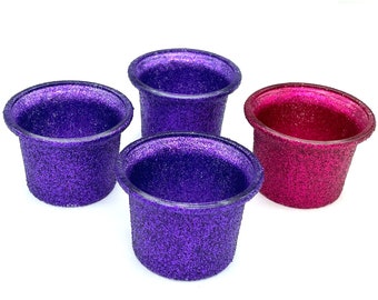 Könnte beinhalten: Vier kleine, mit Glitzer bedeckte Behälter. Drei sind tiefviolett und einer leuchtend magenta. Die Behälter haben eine einfache, abgerundete Form und sind vor einem weißen Hintergrund platziert. Sie scheinen dekorative Gegenstände zu sein.