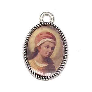 May include: Oval pendant featuring a portrait of a woman in a pink headscarf. The portrait is framed by a silver-coloured, twisted rope-like border. A small loop is at the top for attaching to a chain or bracelet.