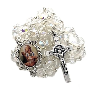 Puede incluir: Un rosario de cristal blanco con un crucifijo de plata y una imagen redonda de un santo. El crucifijo tiene la inscripción "INRI" en el travesaño.