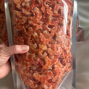 May include: A clear plastic bag filled with dried shrimp. The shrimp are a reddish-orange color and appear to be small and curled. The bag is sealed and held by a hand.