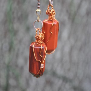 May include: Two orange and brown gemstone pendants hanging from a brass chain. The pendants are wrapped in copper wire.