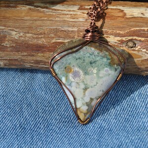 May include: A copper wire wrapped pendant featuring a green, white and brown agate stone with a unique pattern. The pendant is hanging from a copper chain.
