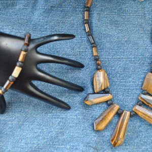 Puede incluir: Un collar y una pulsera de cuentas marrones y negras con piedras de ojo de tigre. El collar tiene un colgante con cinco piedras de ojo de tigre.