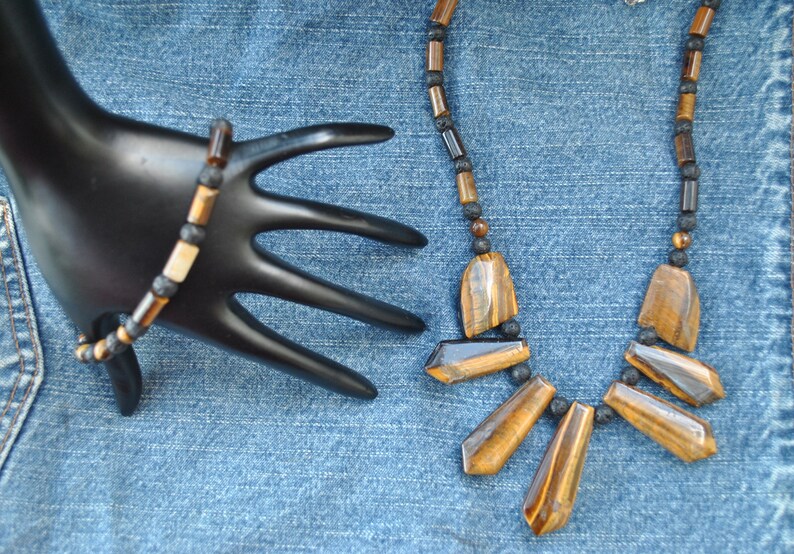 Puede incluir: Un collar y una pulsera de cuentas marrones y negras con piedras de ojo de tigre. El collar tiene un colgante con cinco piedras de ojo de tigre.