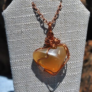 May include: A heart-shaped pendant necklace with a copper wire wrap. The pendant is a translucent, amber-colored stone. The necklace is displayed on a gray fabric jewelry stand.
