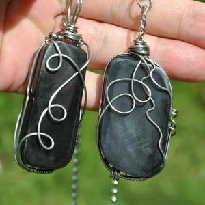 May include: Two rectangular, black stone pendants with silver wire wrapping and chain. The wire is intricately wrapped around the stones in a decorative pattern. The pendants are suspended from a silver chain.