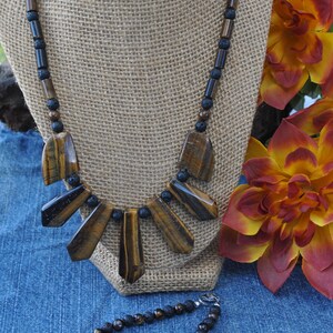 Puede incluir: Un collar de cuentas marrones y negras con un colgante de ojo de tigre. El collar se muestra en un maniqu&iacute; de arpillera. Tambi&eacute;n se muestra una pulsera a juego de cuentas marrones y negras.