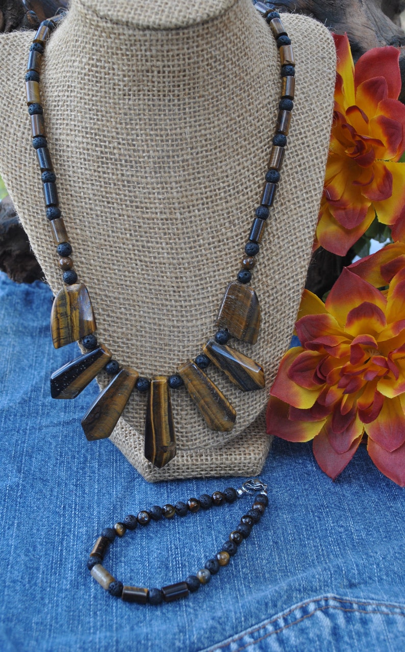 Puede incluir: Un collar de cuentas marrones y negras con un colgante de ojo de tigre. El collar se muestra en un maniqu&iacute; de arpillera. Tambi&eacute;n se muestra una pulsera a juego de cuentas marrones y negras.