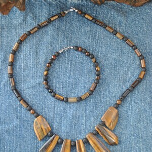 Puede incluir: Un conjunto de dos collares hechos con cuentas de ojo de tigre y roca de lava negra. El collar m&aacute;s grande tiene un colgante con cinco piedras de ojo de tigre en forma de abanico. El collar m&aacute;s peque&ntilde;o es un brazalete.