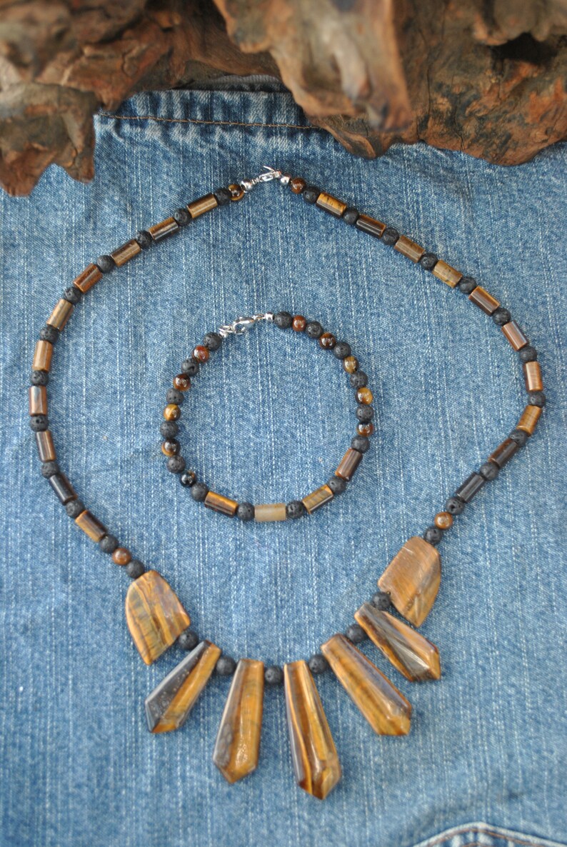 Puede incluir: Un conjunto de dos collares hechos con cuentas de ojo de tigre y roca de lava negra. El collar m&aacute;s grande tiene un colgante con cinco piedras de ojo de tigre en forma de abanico. El collar m&aacute;s peque&ntilde;o es un brazalete.
