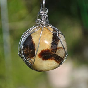 May include: A pendant featuring a polished, spherical stone with a cream and brown pattern, encased in a silver wire cage. The pendant hangs from a silver ring, suitable for a keychain or necklace.