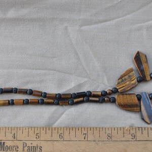 Puede incluir: Un collar de cuentas marrones y negras con un cierre plateado. El collar presenta un gran colgante en forma de abanico hecho de piedras de ojo de tigre.