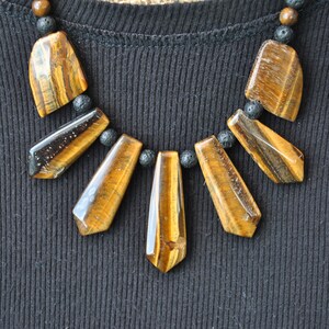 Puede incluir: Un collar con un colgante de ojo de tigre marr&oacute;n y dorado y cuentas de lava negra.