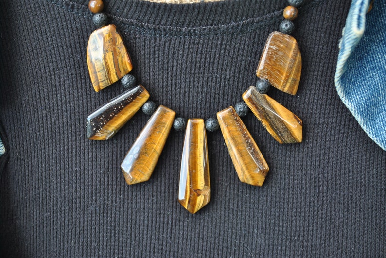 Puede incluir: Un collar con un colgante de ojo de tigre marr&oacute;n y dorado y cuentas de lava negra.