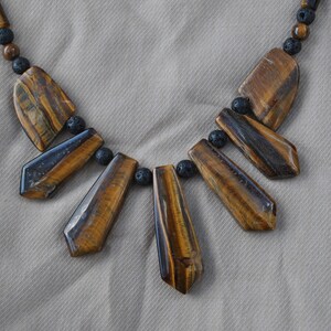 Puede incluir: Un collar de piedra de ojo de tigre marr&oacute;n y dorado con cuentas de lava negra. El collar presenta seis piedras de ojo de tigre en forma de l&aacute;grima.