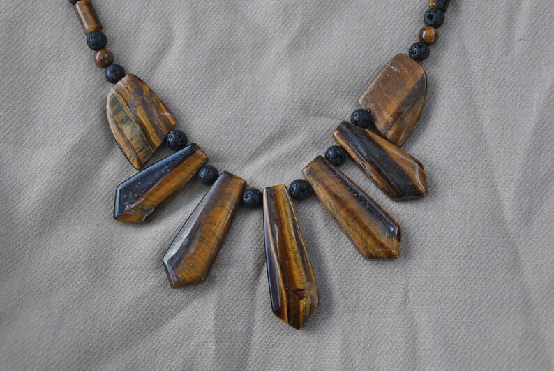 Puede incluir: Un collar de piedra de ojo de tigre marr&oacute;n y dorado con cuentas de lava negra. El collar presenta seis piedras de ojo de tigre en forma de l&aacute;grima.