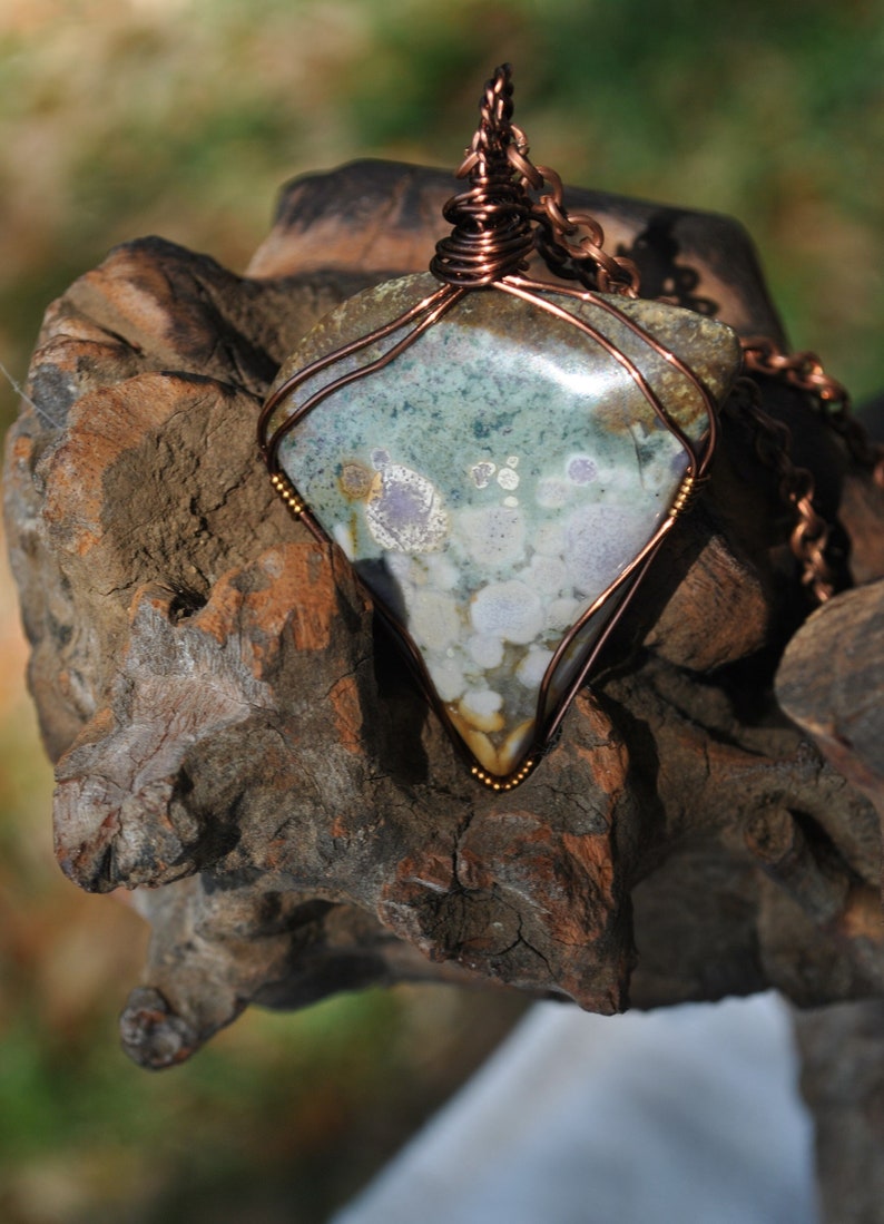 May include: A copper wire wrapped pendant featuring a green, white and brown stone with a unique pattern. The pendant is hanging from a copper chain.