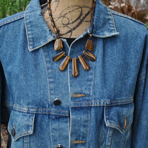 Puede incluir: Una chaqueta vaquera con un collar de piedra de ojo de tigre marr&oacute;n y negro. El collar tiene un dise&ntilde;o en forma de abanico con varias piedras.