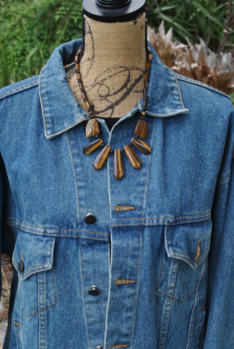 Puede incluir: Una chaqueta vaquera con un collar de piedra de ojo de tigre marr&oacute;n y negro. El collar tiene un dise&ntilde;o en forma de abanico con varias piedras.