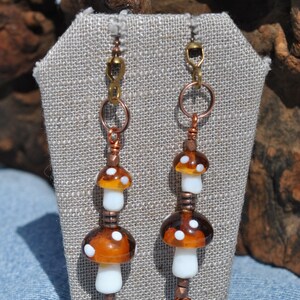May include: A pair of dangle earrings featuring two stacked glass mushroom beads on each side. The mushrooms have white stems and translucent brown caps with white spots. The beads are connected by copper-toned wire, rings, and small metallic spacers.