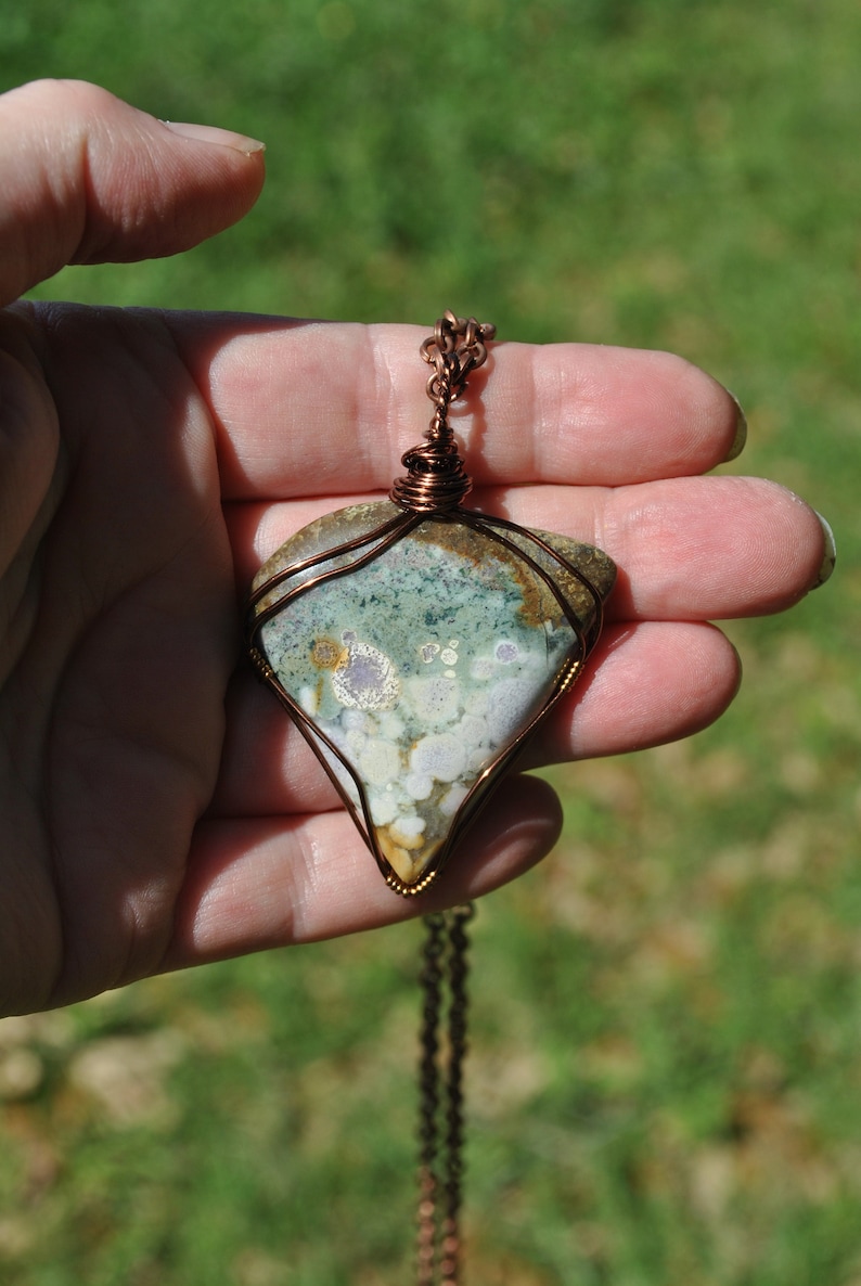 May include: A copper wire wrapped pendant featuring a green, white and brown agate stone with a unique pattern. The pendant is attached to a copper chain.