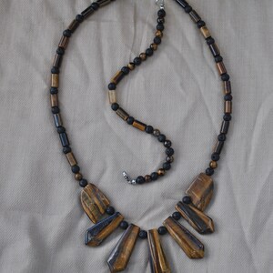 Puede incluir: Un collar de cuentas marrones y negras con un colgante de ojo de tigre. El colgante est&aacute; hecho de varias piezas largas y puntiagudas de piedra de ojo de tigre.