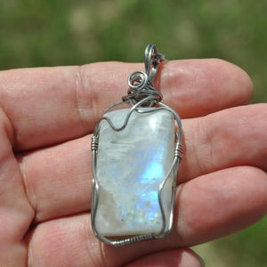 May include: A rectangular moonstone pendant featuring a smooth, translucent white surface with iridescent blue flashes. The gemstone is secured in a silver-toned wire-wrapped setting with a coiled loop at the top for hanging on a chain or cord.
