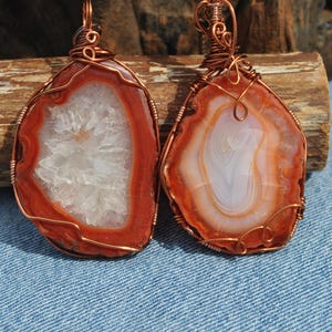 May include: Two pendants featuring irregular, polished agate slices with deep orange and rust-colored outer bands. One pendant displays a central white crystalline geode cavity, while the other shows concentric white and orange banding, both wrapped in copper wire.