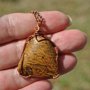 May include: A handcrafted pendant featuring a triangular, patterned stone in shades of brown and yellow. The stone is wrapped in copper wire and suspended from a matching chain. The pendant is held in a hand against a blurred background.