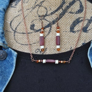 May include: A copper chain necklace with a brown and white bead bar pendant and matching earrings. The necklace and earrings feature brown, white, and amber beads.