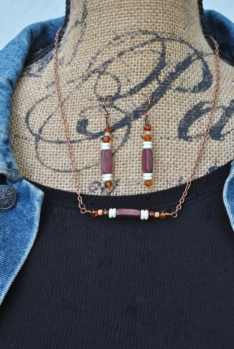 May include: A copper chain necklace with a brown and white bead bar pendant and matching earrings. The necklace and earrings feature brown, white, and amber beads.