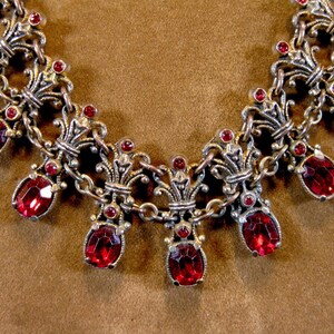 May include: Close-up of a silver-toned necklace with ornate filigree details. The necklace features large, oval-shaped, red gemstones and smaller red accent stones. The chain is composed of small, linked segments.