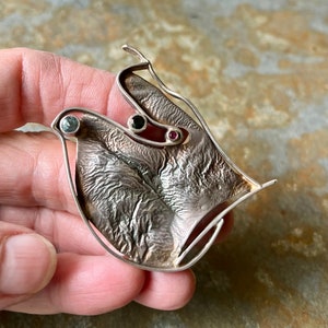 May include: A silver pendant with a textured, abstract design. The pendant features three gemstones: a green one, a black one, and a pink one.