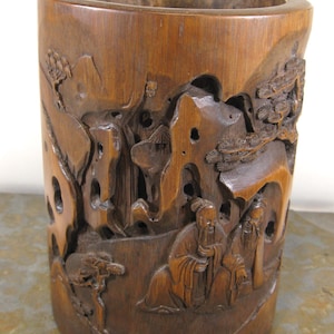 May include: A cylindrical wooden container with intricate relief carvings. The carvings depict a landscape with figures, trees, and mountains. The container is made of a warm brown wood and has a smooth, polished finish.