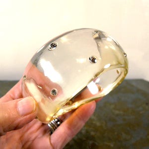 May include: A wide, clear, translucent resin bangle bracelet featuring several small, round, embedded silver-toned crystals scattered across the smooth, curved surface of the circular accessory.